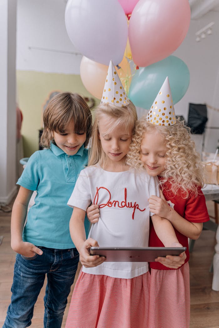 Three children enjoying a virtual birthday celebration with balloons and party hats.